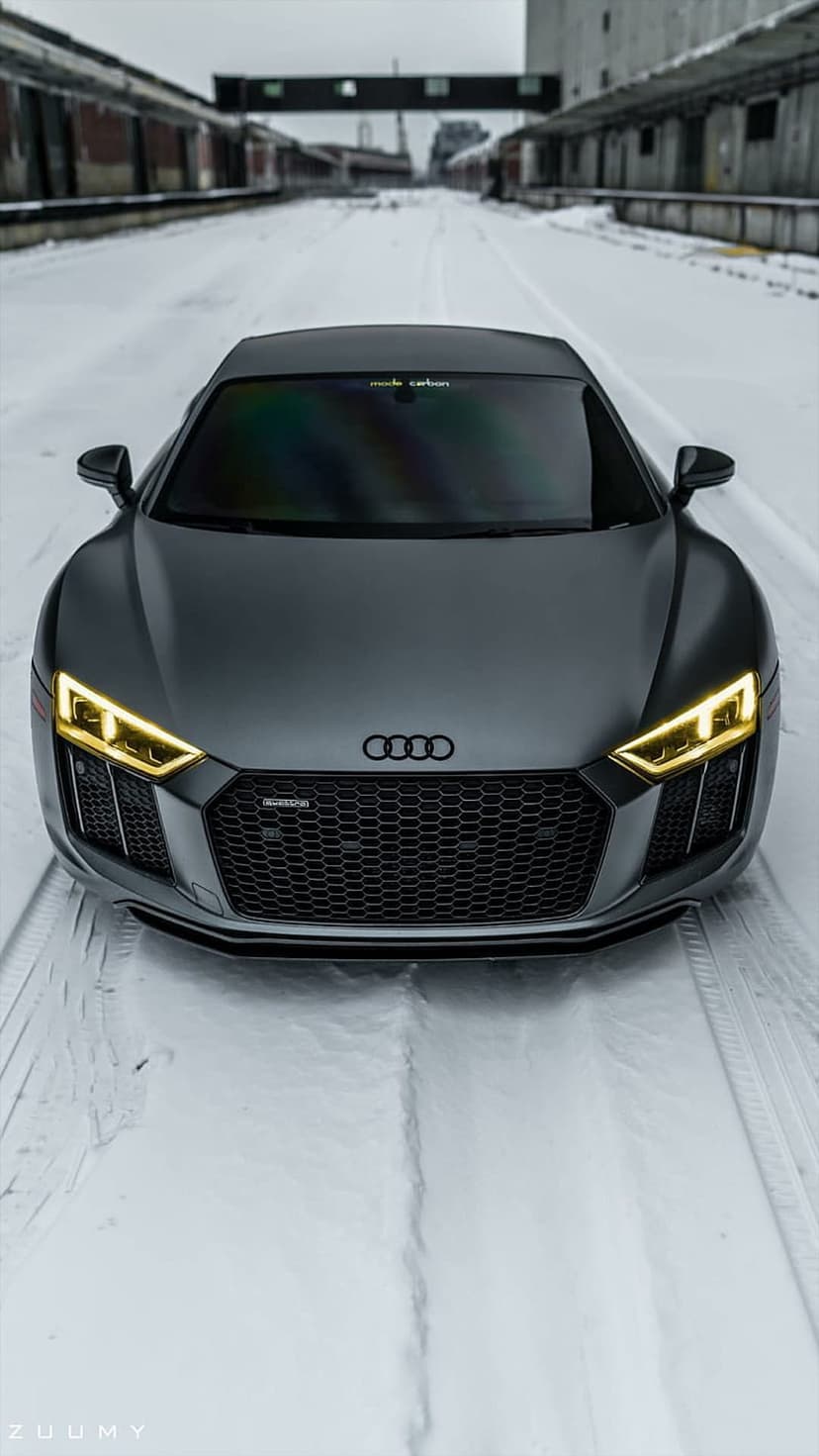 Matte grey Audi R8 supercar parked on a snowy road with glowing yellow LED headlights and a blurred bridge.