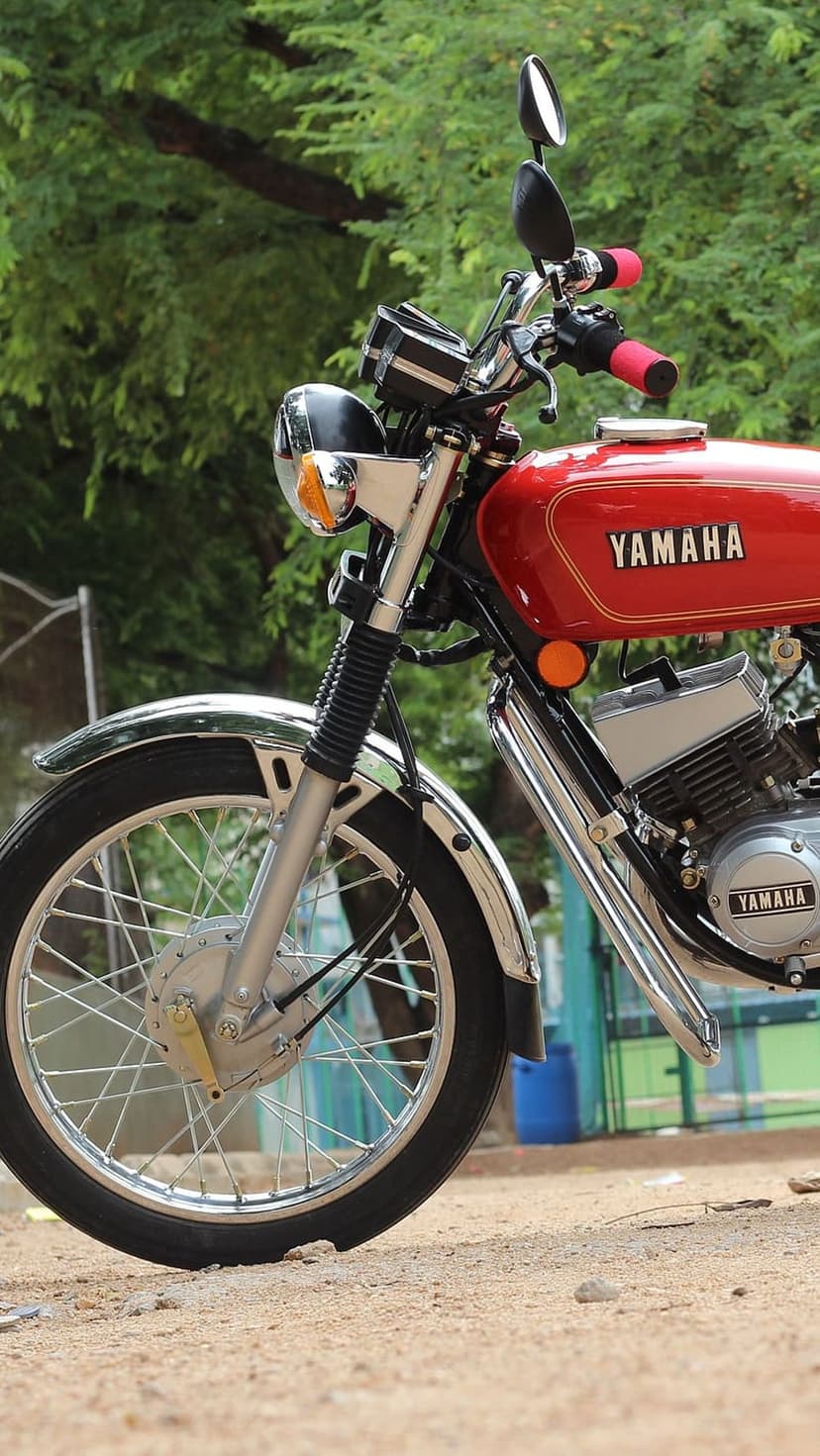 Yamaha RX100 front wheel with a chrome fender, red fuel tank, and spoked rim against a soft green forest.