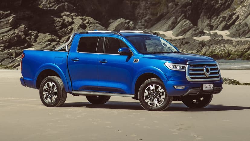 Metallic blue GWM Cannon pickup truck on a wide sandy beach with rocky coastal cliffs in the background.