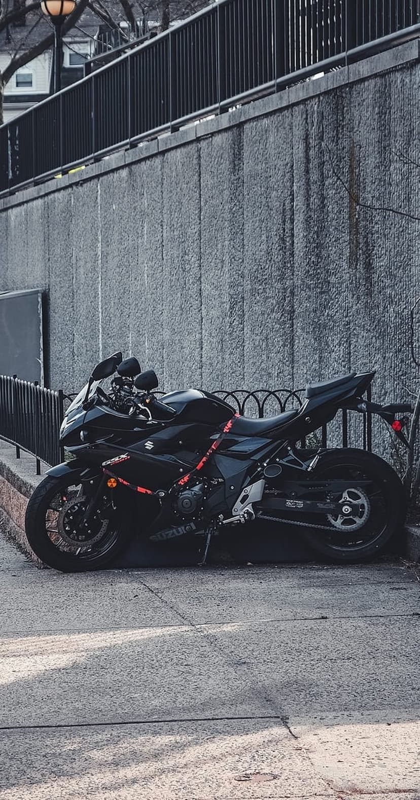 Black Suzuki motorcycle with red accents parked on a sidewalk beside a textured grey concrete wall in sunlight.