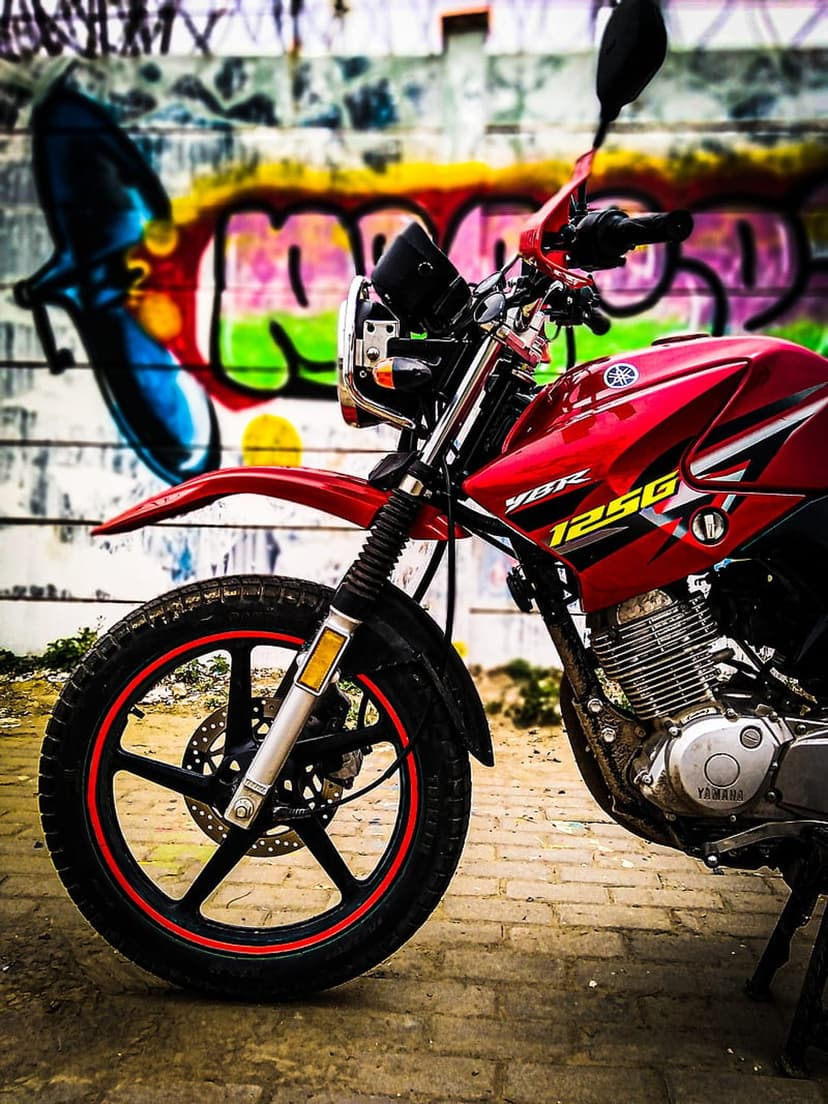 Yamaha 125G motorcycle with red paint and black wheels stands before a colorful graffiti-covered brick wall.