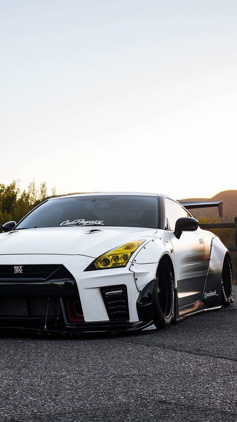 White Nissan GT-R with widebody kit and yellow headlights parked outdoors under a dusky sky at a low angle.