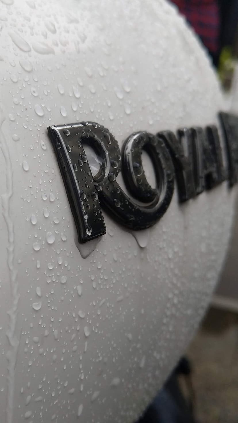 Royal Enfield chrome logo with glistening water droplets on a white motorcycle tank in a vertical close-up.