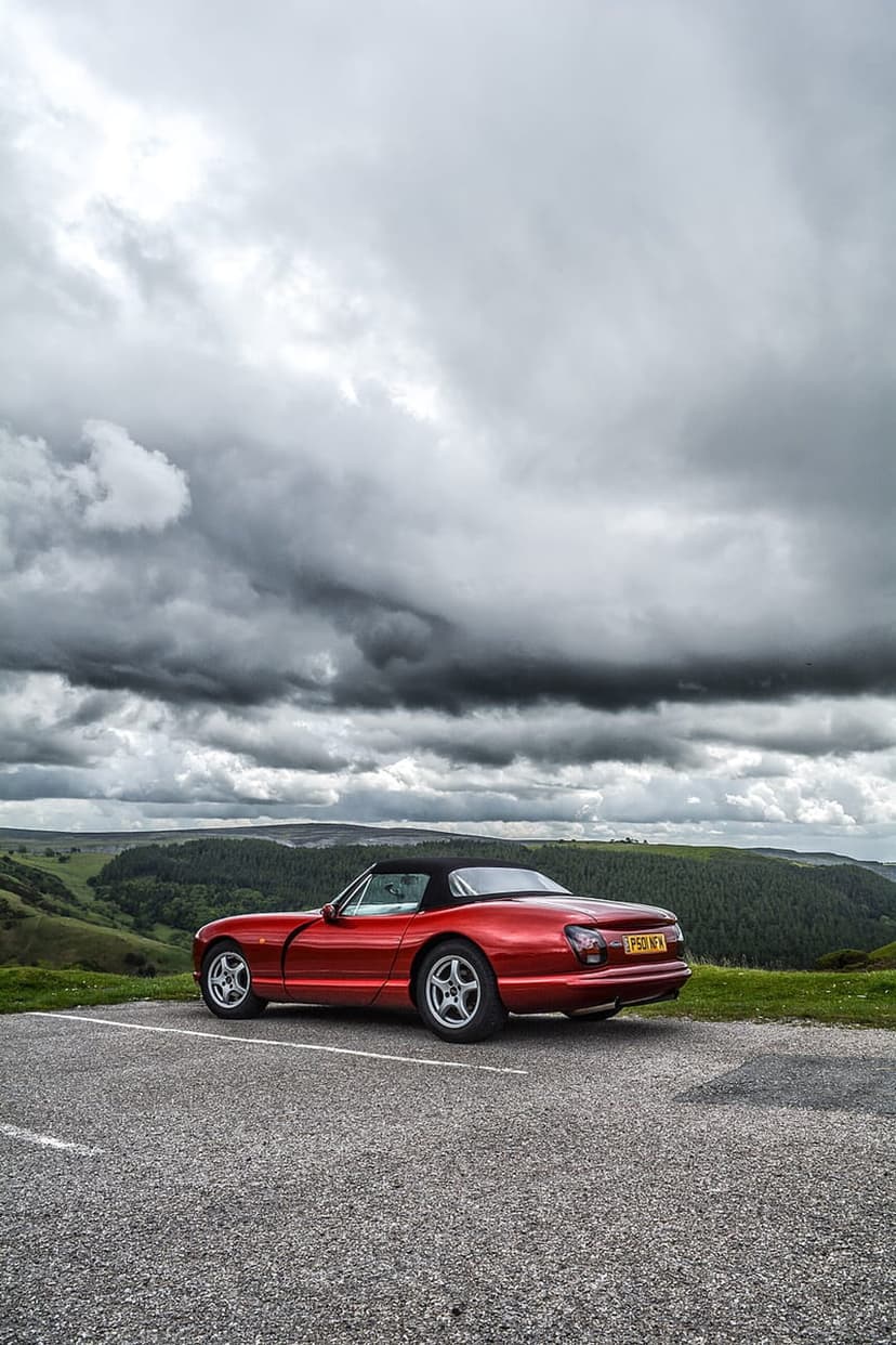 Red convertible sports car parked on gravel overlooking a green valley under dark, dramatic storm clouds.