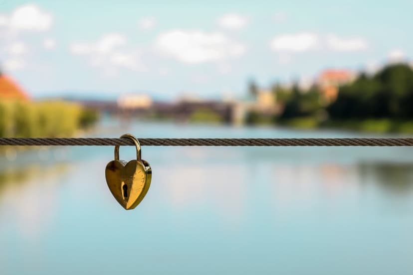 Golden heart-shaped lock secured to a steel bridge cable with a blurred river and cityscape in the background.
