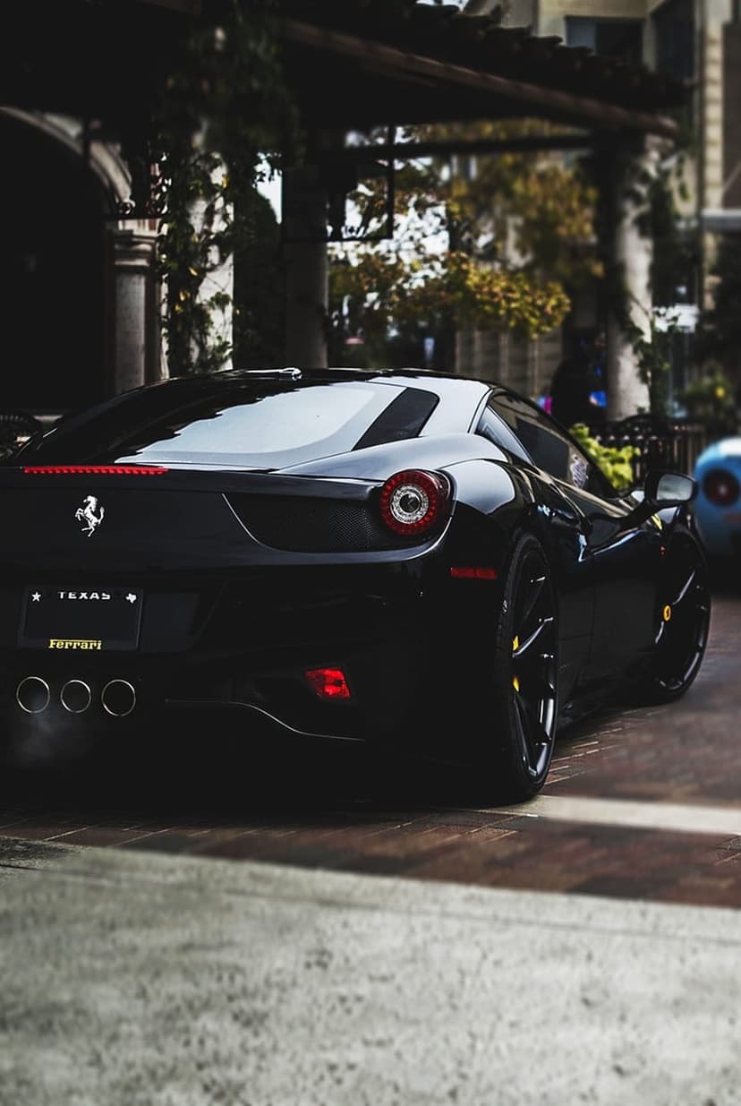 Ferrari 458 Italia in black with Texas plates and quad exhaust pipes parked on a paved road at a low rear angle.