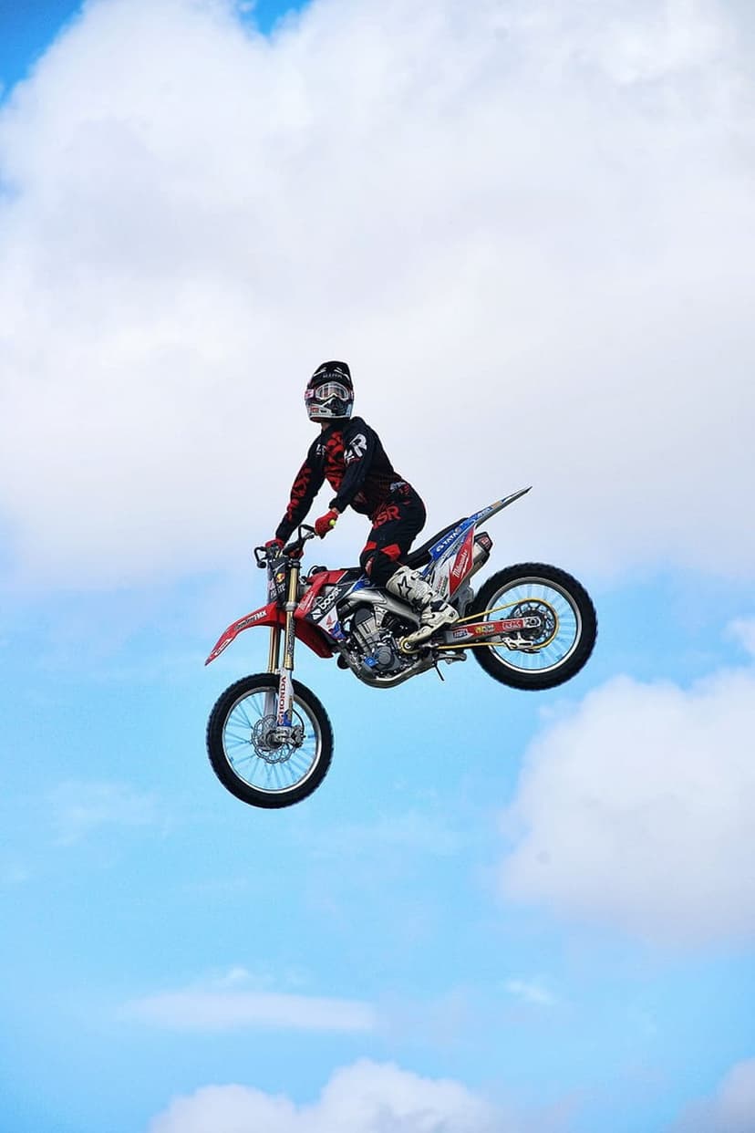 Motocross rider wearing a full helmet jumps a dirt bike mid-air among fluffy white clouds in a blue sky.