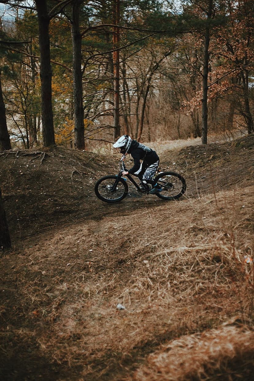 Mountain biker wearing a helmet leans into a sharp turn on a steep dirt path through a dense pine forest.