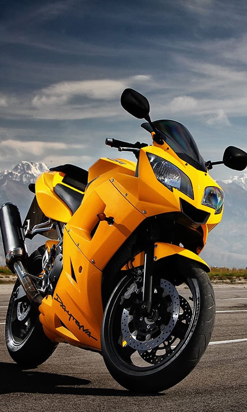 Yellow Daytona motorcycle parked on a road with a scenic mountain range and cloudy sky in the background.