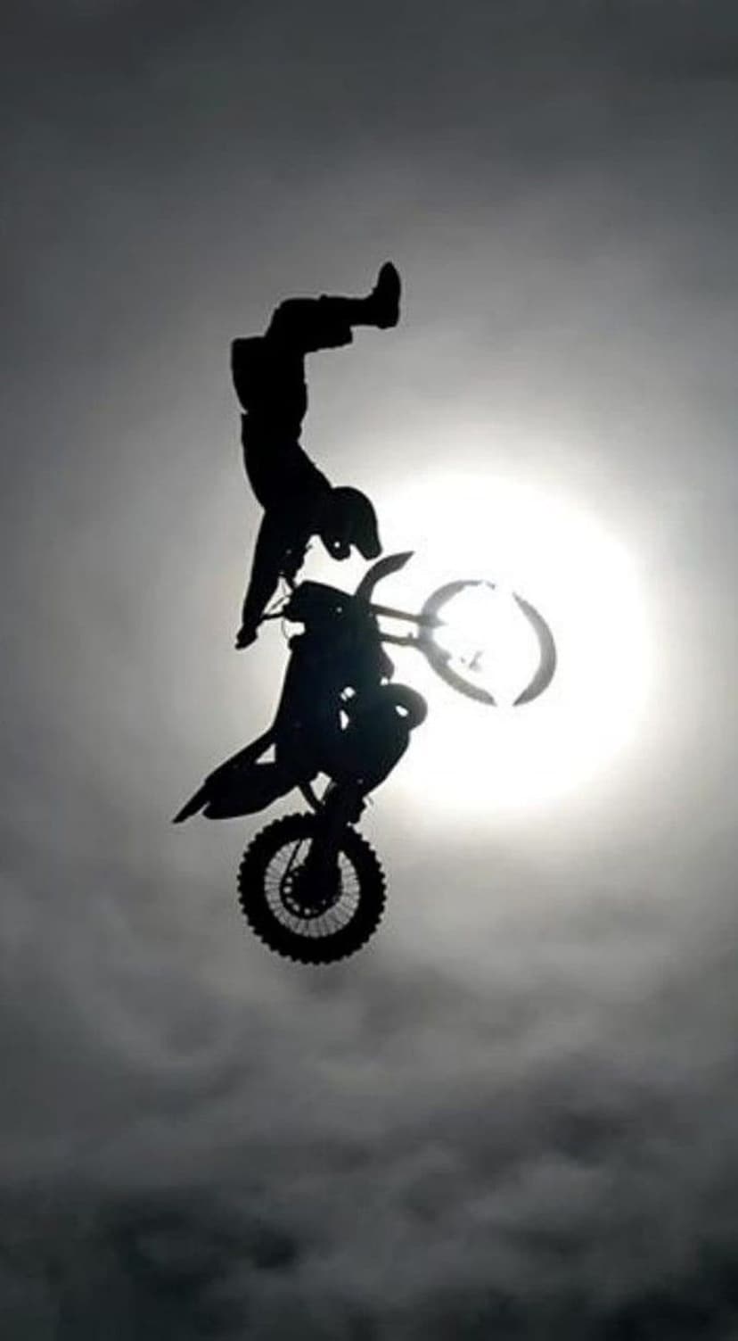 Motocross rider performing a mid-air handstand stunt as a dark silhouette against a bright, hazy sun and clouds.
