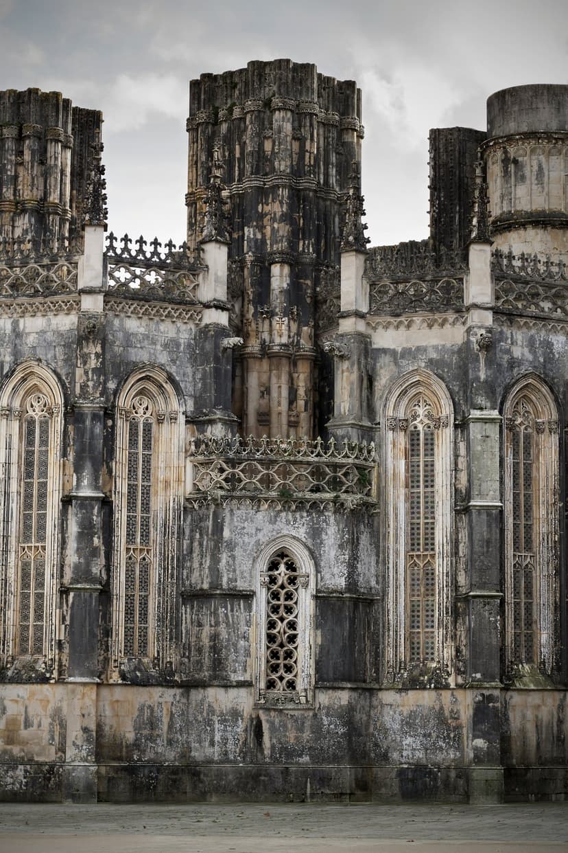 Gothic arched windows and ornate stone carvings of Batalha Monastery stand against a dramatic cloudy gray sky.