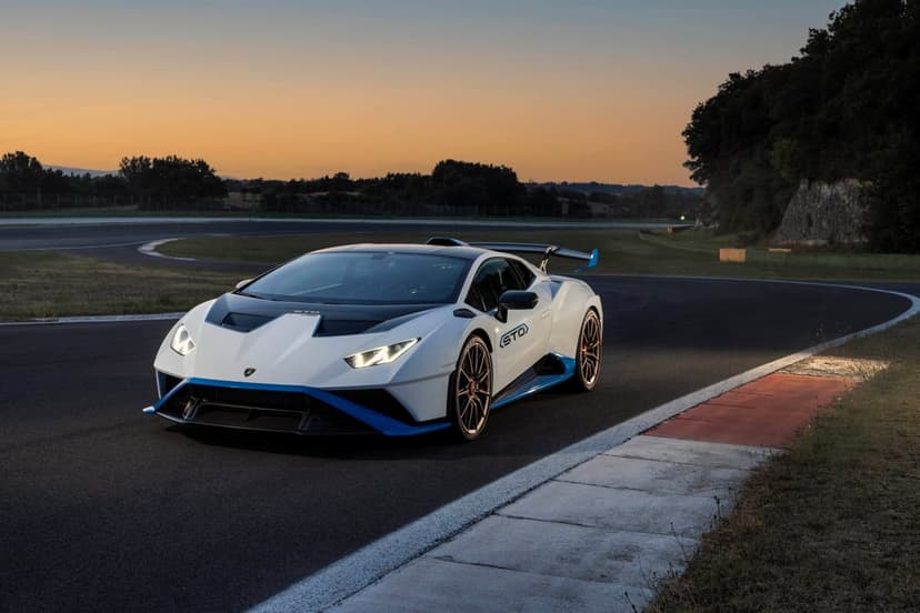 White Lamborghini Huracán STO with blue accents parked on an asphalt race track curve during a golden sunset.