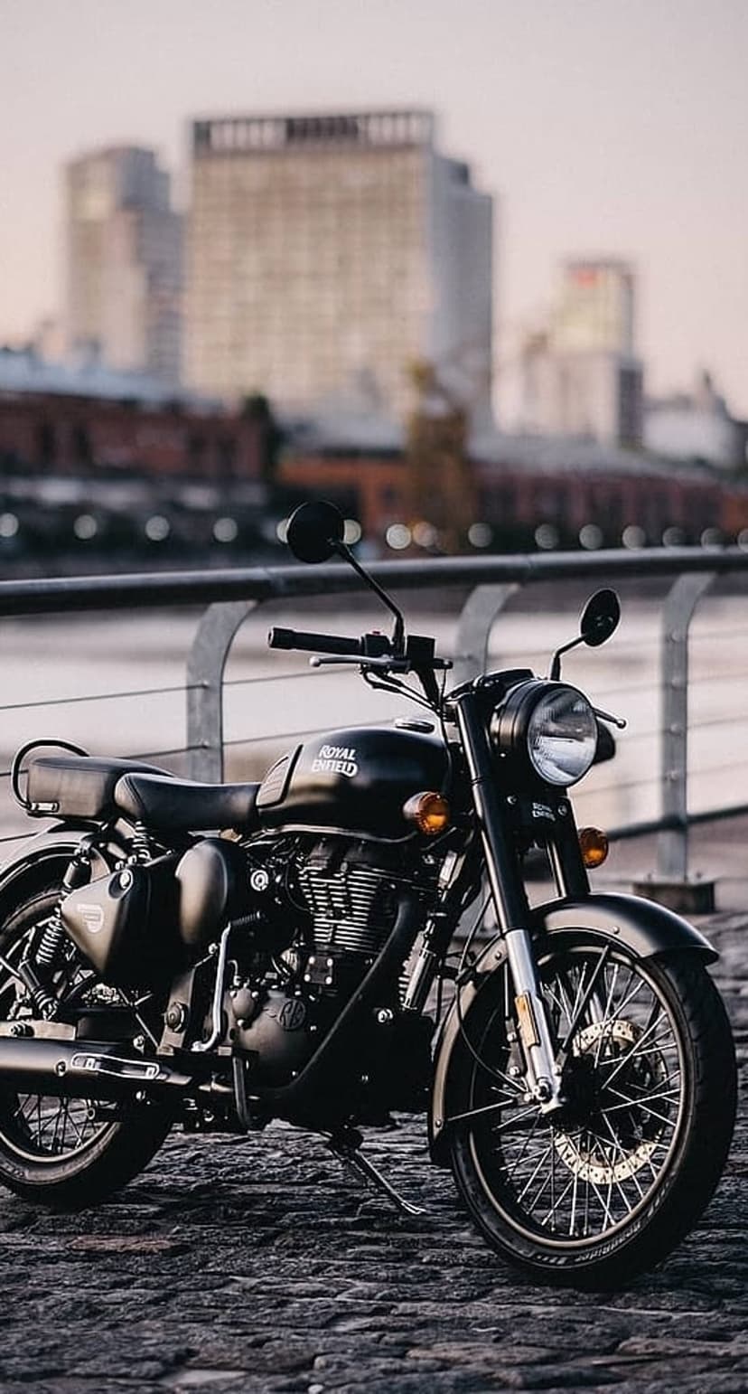 Black Royal Enfield motorcycle parked on cobblestones with a blurred urban waterfront and buildings behind it.