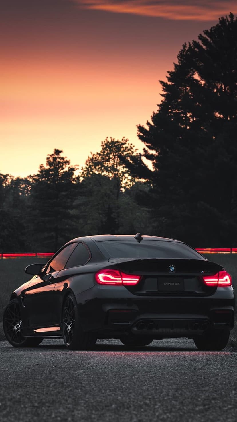 Black BMW M4 parked on a gravel road with glowing red taillights under an orange and pink sunset sky.