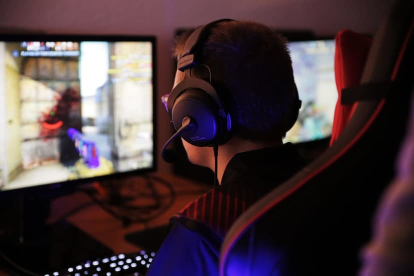 Gamer from behind wearing black headphones playing an FPS video game in a room with blue and red neon lighting.