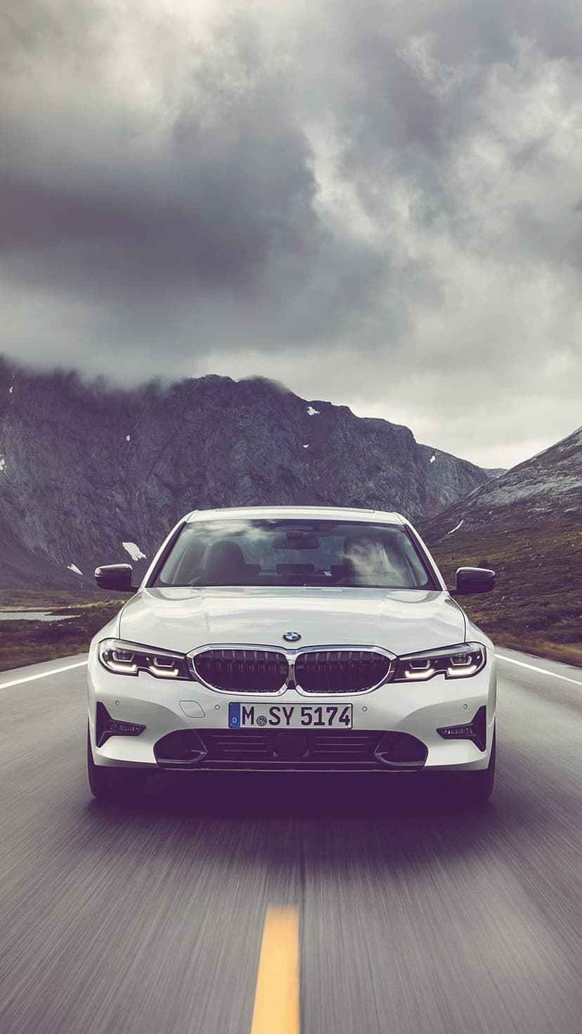 White BMW sedan with illuminated headlights driving on a mountain road beneath a cloudy sky with snowy peaks.