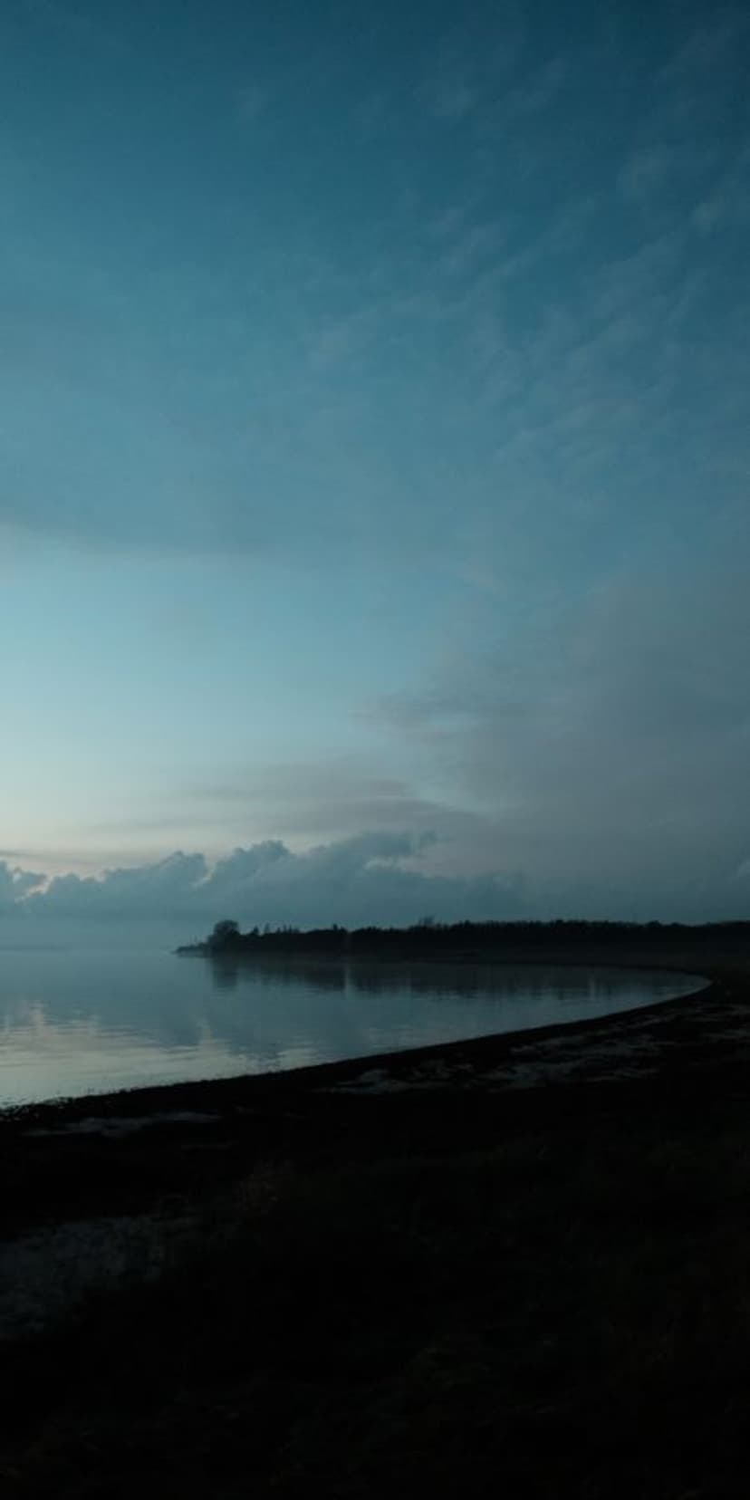 Misty coastline at dawn with calm water reflecting a soft blue sky and a dark silhouetted shore in the foreground.