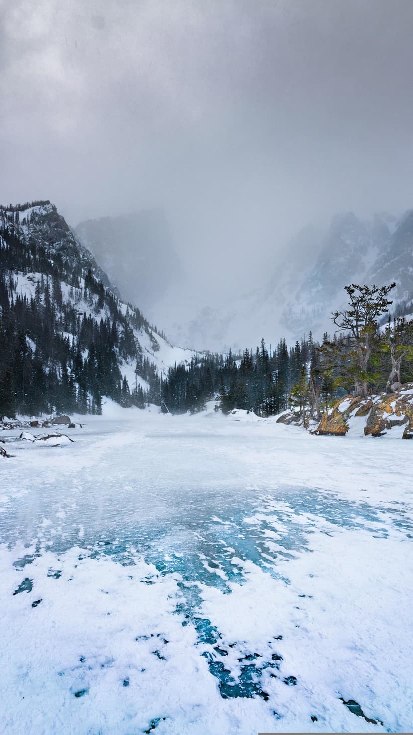 Frozen glacial lake with blue ice shards sits before towering snowy mountains and evergreen forests.