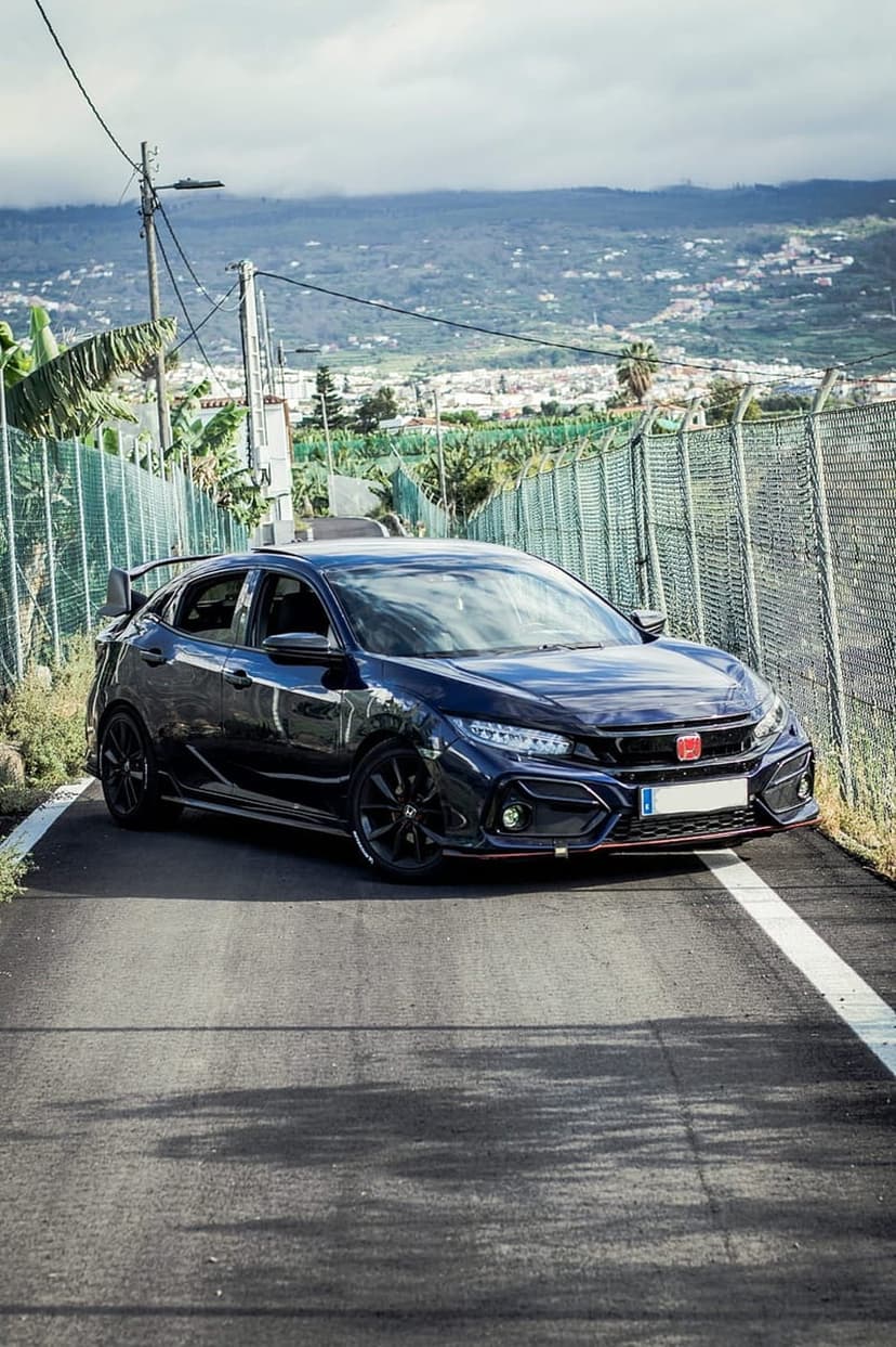 Dark blue Honda Civic Type R with a rear spoiler and black wheels on a paved road against a green hillside.