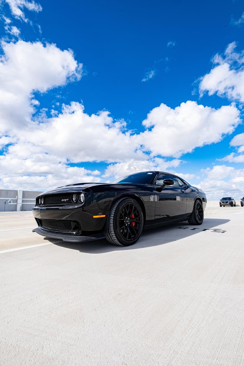 Black Dodge Challenger SRT Hellcat on a rooftop with red calipers, alloy wheels, and a bright blue cloudy sky.