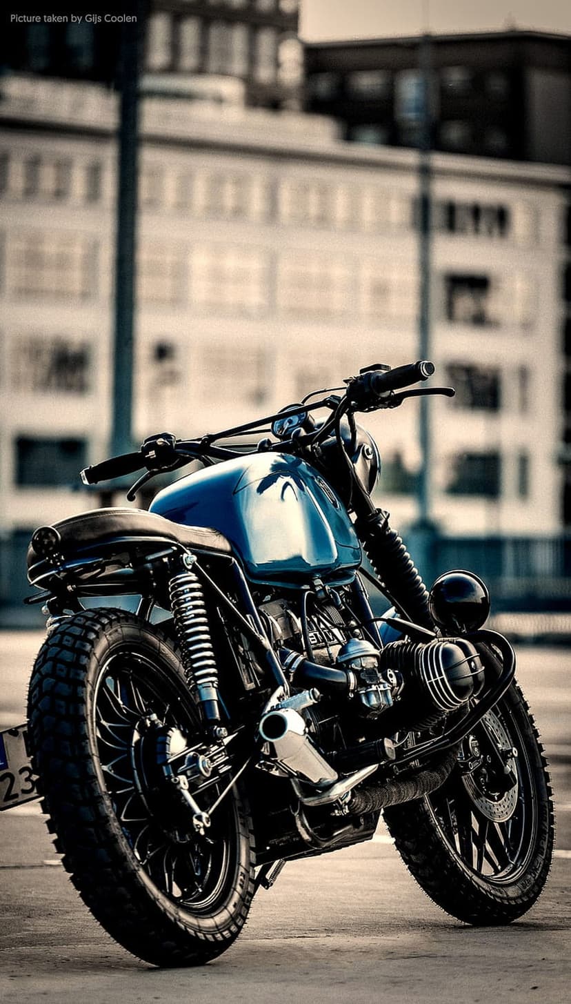 Blue cafe racer motorcycle with a chrome fuel tank and black seat parked on a blurred urban street background.