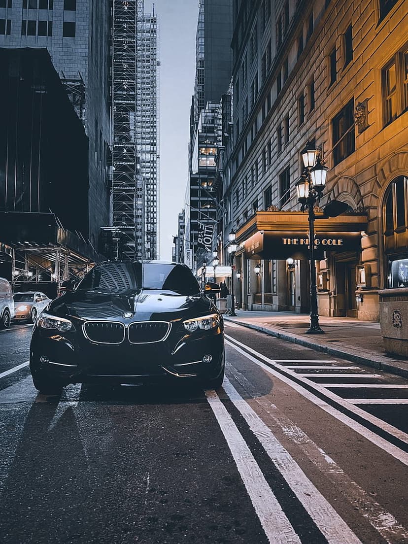 Black luxury car parked on wet asphalt at night, surrounded by illuminated skyscrapers and city buildings.