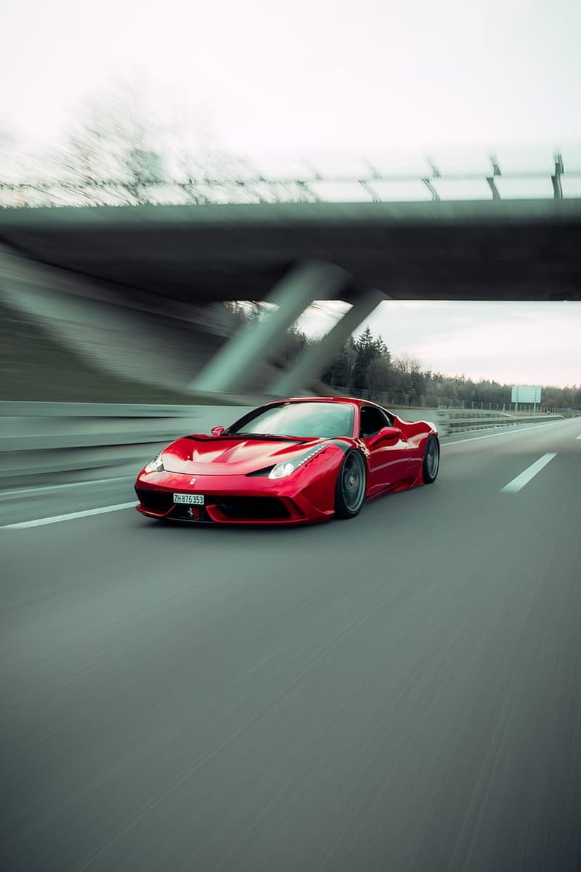 Red Ferrari 458 Italia supercar driving fast on a highway under a concrete bridge with motion-blurred trees.