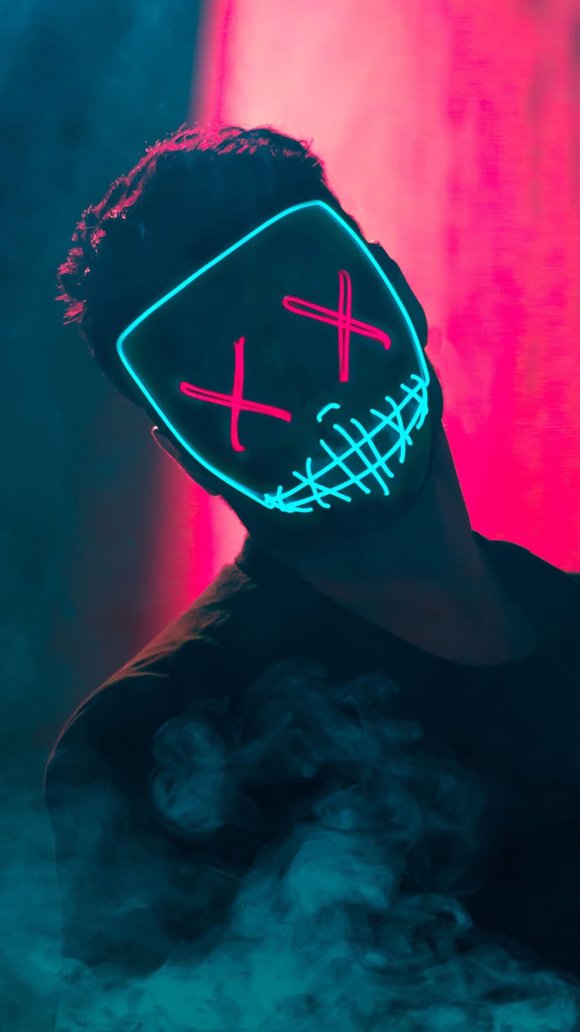 Glowing mask with pink X-shaped eyes and a blue jagged grin emerges from smoke against a blurred background.