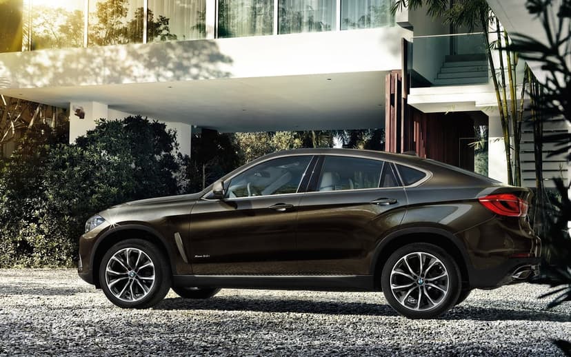 Bronze BMW X6 parked on a gravel driveway in front of a modern white house with golden sunlight flares.