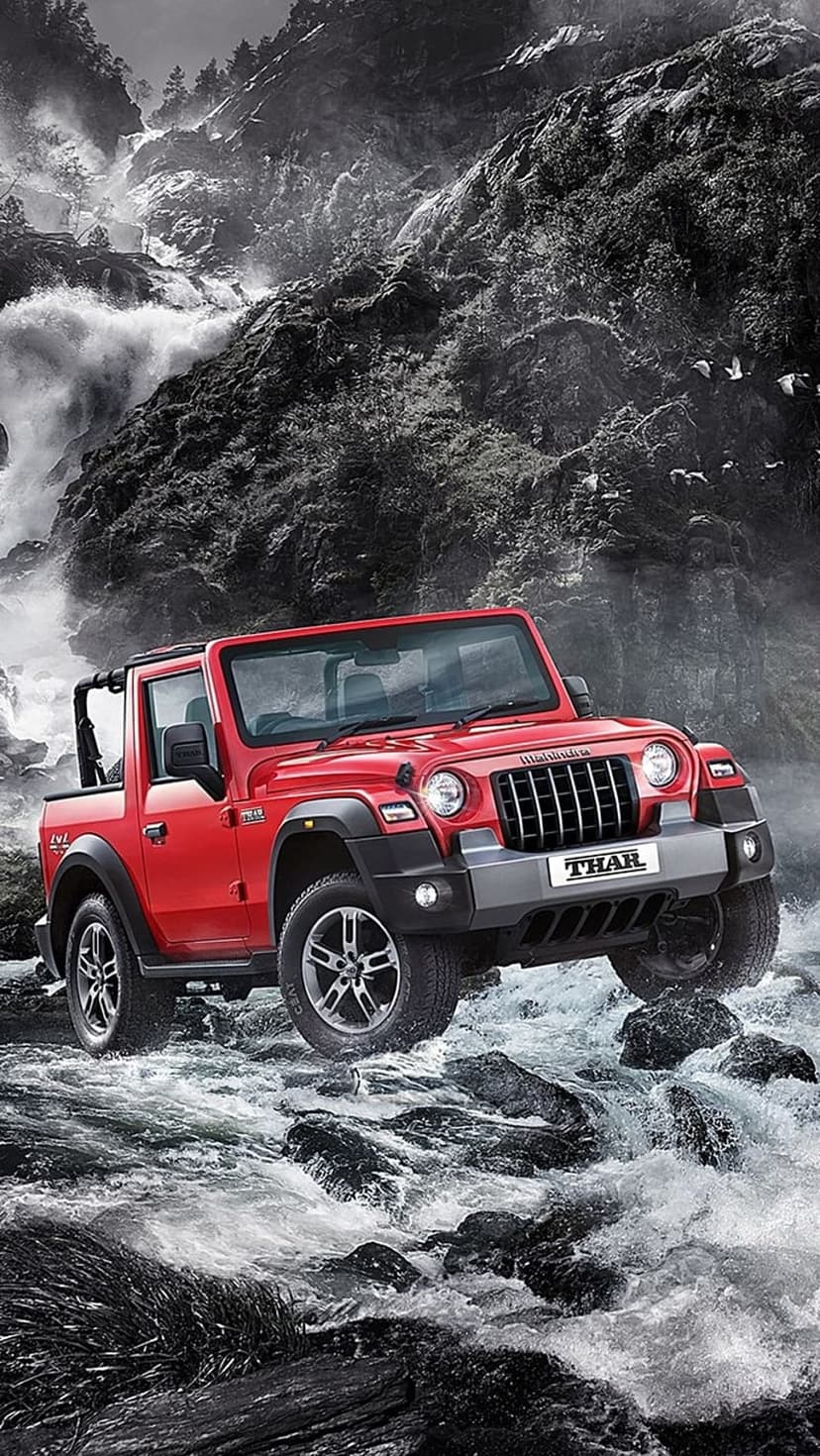 Red Mahindra Thar SUV drives through splashing river water over dark grey rocks with a mountain background.
