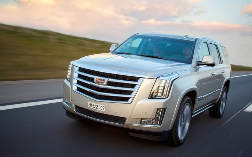 Silver Cadillac Escalade SUV driving on a highway with motion blur against a sunset sky and green hills.