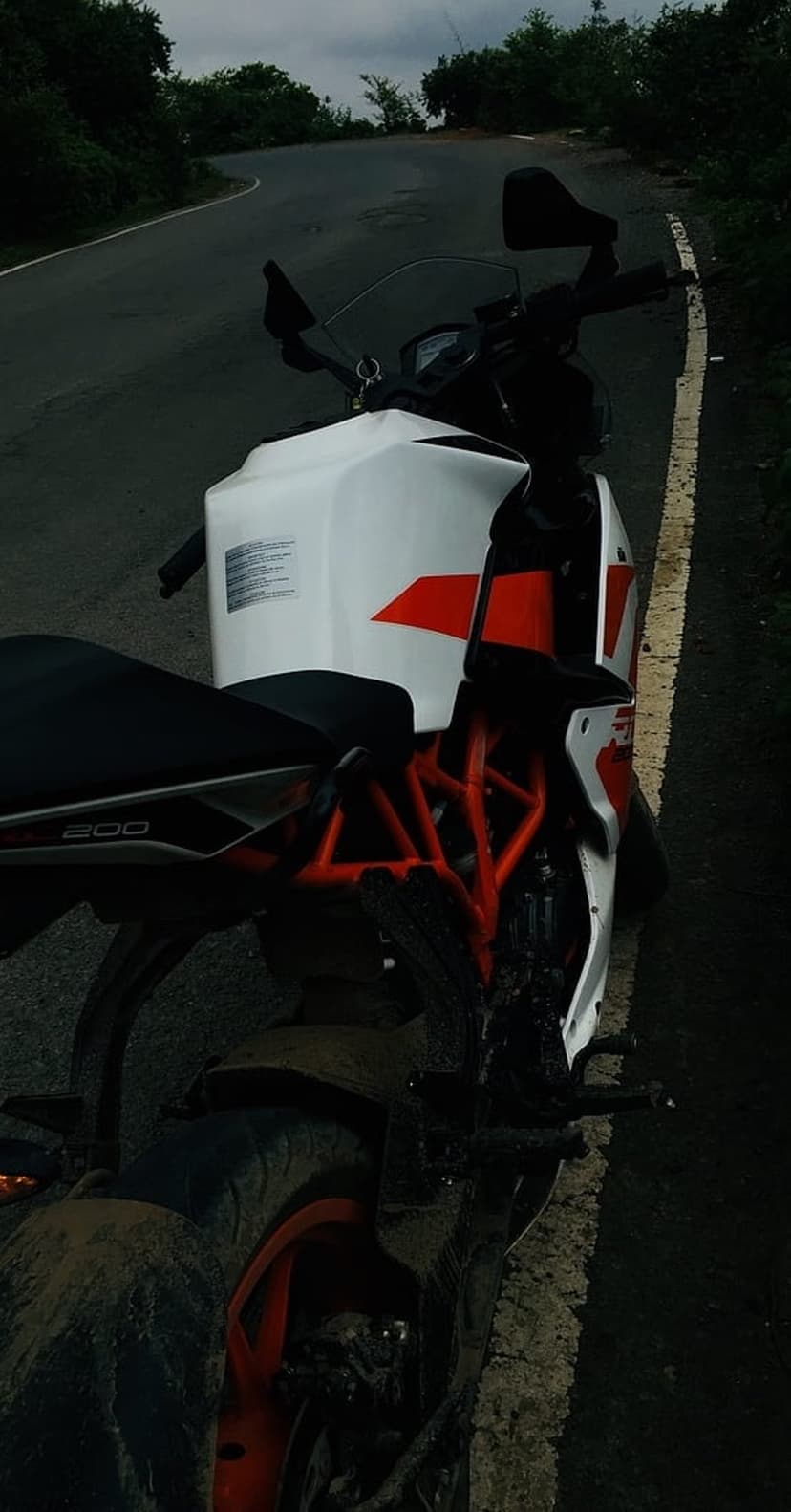 KTM RC 200 motorcycle with an orange frame parked on a wet, winding asphalt road under a cloudy, overcast sky.
