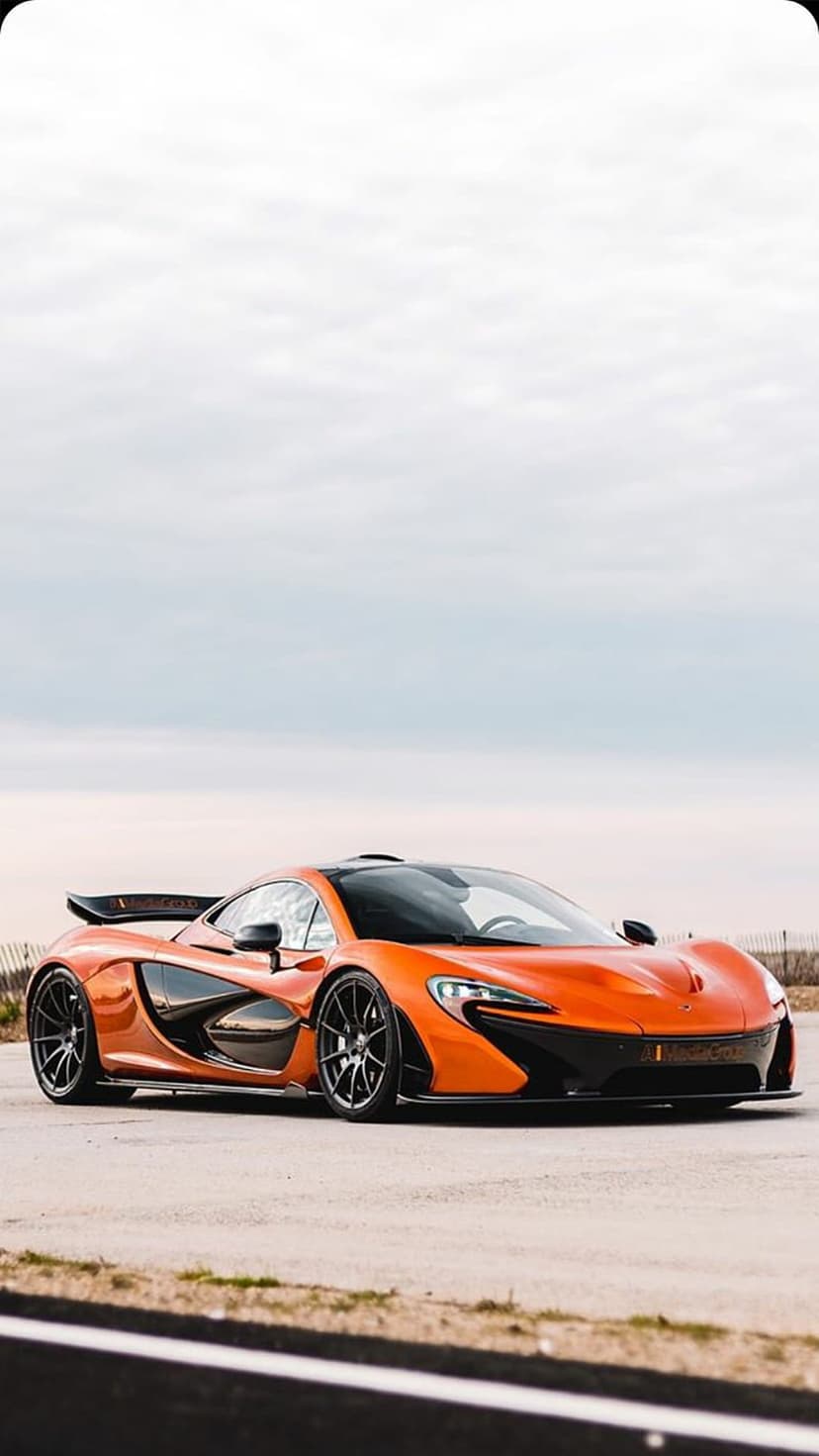 Orange McLaren P1 supercar with black wheels on a paved road under a grey overcast sky at a low camera angle.