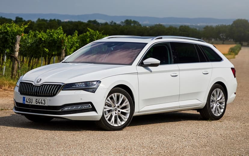 White Skoda Superb Combi wagon on a gravel road with green vineyards and rolling hills in the background.