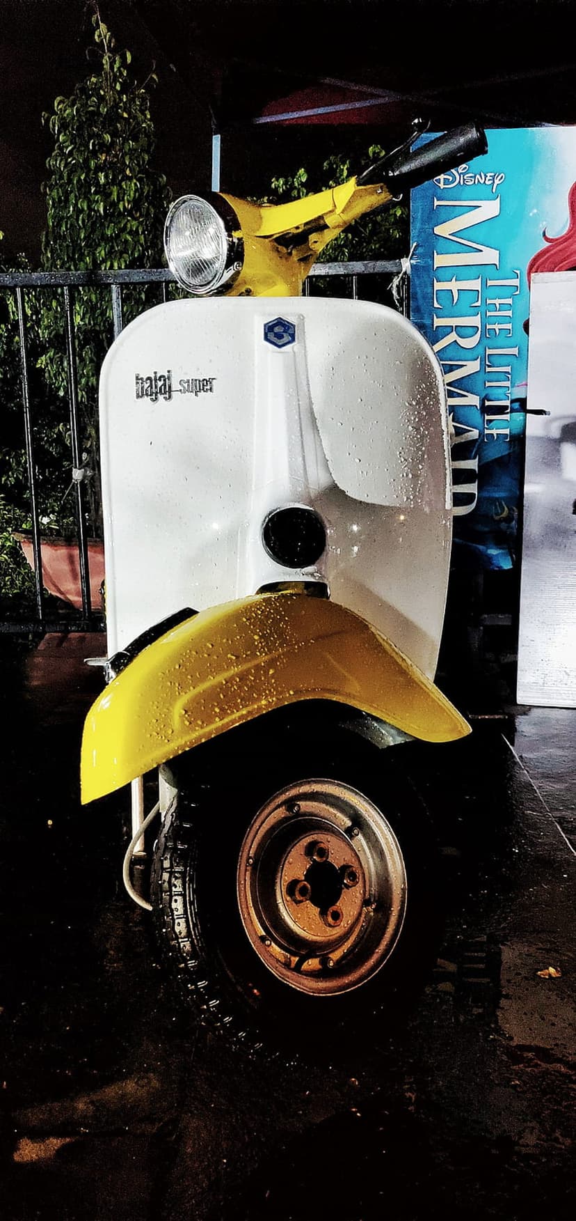 Yellow and white Bajaj Super scooter parked on wet ground at night with a blue Little Mermaid banner behind it.