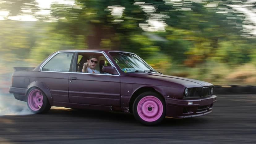 Purple BMW E30 coupe drifts on a track with vibrant pink wheels and thick white smoke from the rear tires.