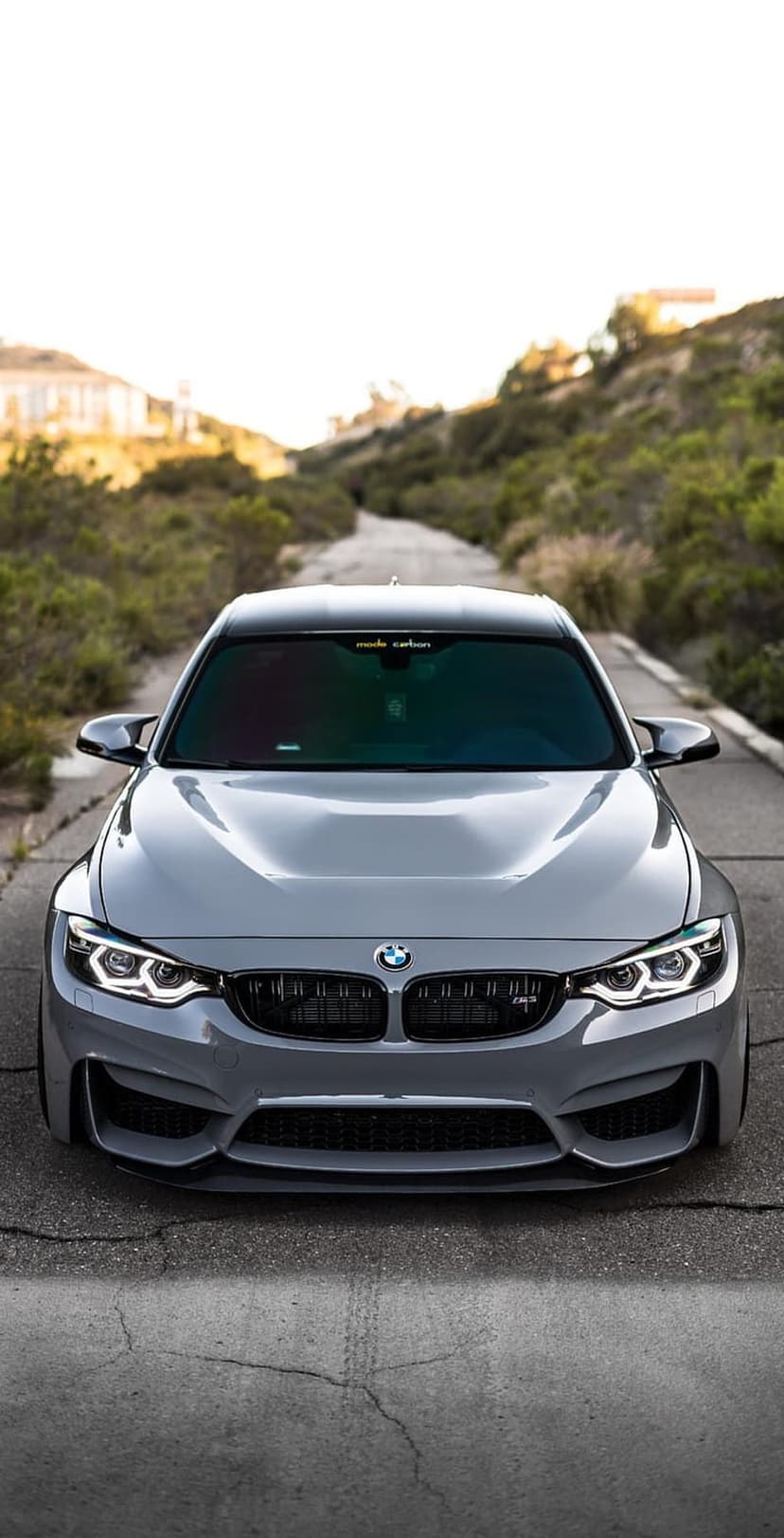 BMW M4 in metallic grey parked on a cracked asphalt road with green rolling hills under a bright clear sky.