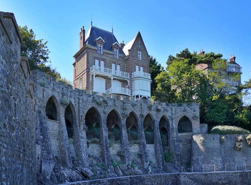 Gothic stone arches support a grand brick villa with white balconies under a clear blue sky with a lone pedestrian.