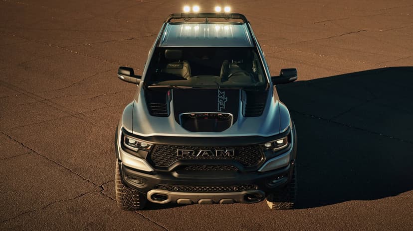 Silver Ram 1500 TRX pickup truck with glowing roof lights and black hood graphics on textured asphalt.