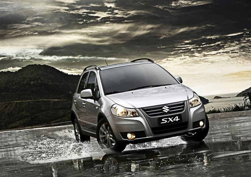 Silver Suzuki SX4 splashing through a puddle on a wet road under a dramatic cloudy sky and coastal hills.