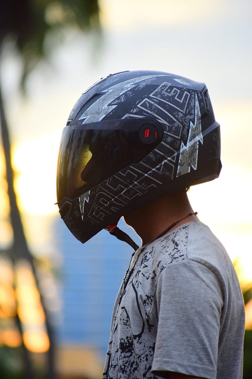 Man wearing a black motorcycle helmet with silver graphics against a sunset sky and blurred palm trees.