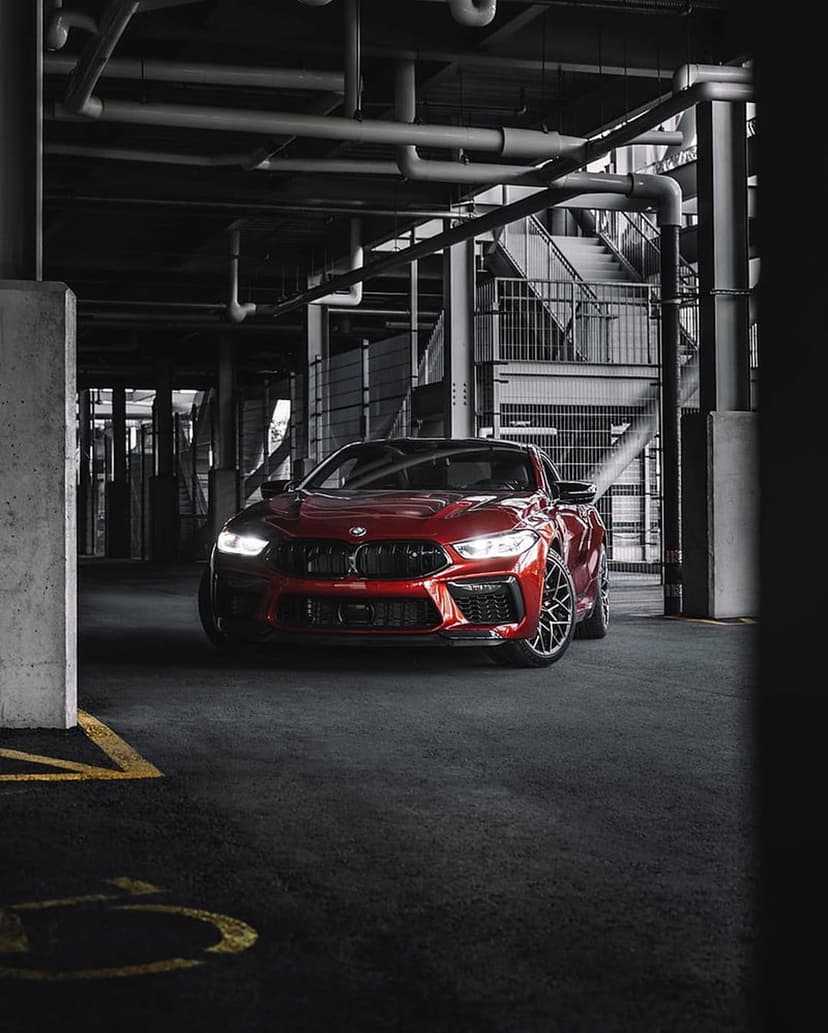 Red BMW M8 parked in a dark industrial garage with exposed pipes, focusing on the car's aggressive front lines.