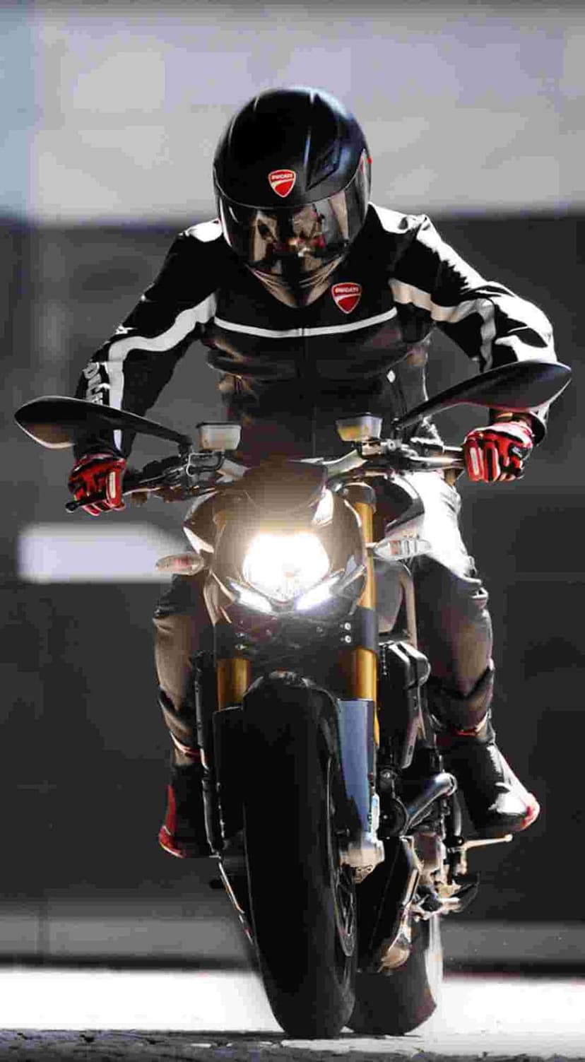 Ducati rider in a black suit and helmet grips handlebars with red gloves near a glowing motorcycle headlight.