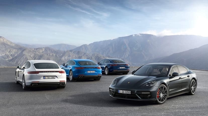 Four Porsche Panamera sedans parked on a mountain road overlooking misty peaks under a clear blue sky.