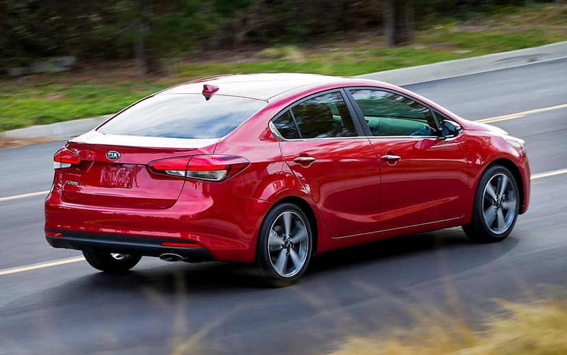 Red Kia Forte sedan driving along a highway with visible motion blur and lush green trees in the background.