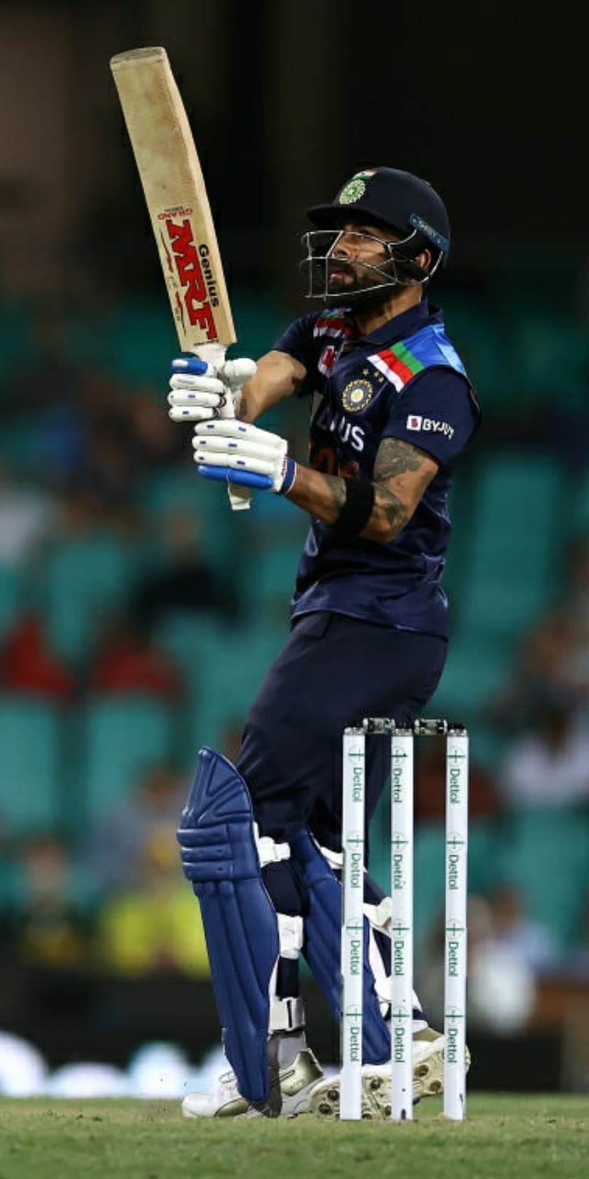 Virat Kohli follows through on a cricket shot wearing a blue jersey and helmet in front of stadium seating area.