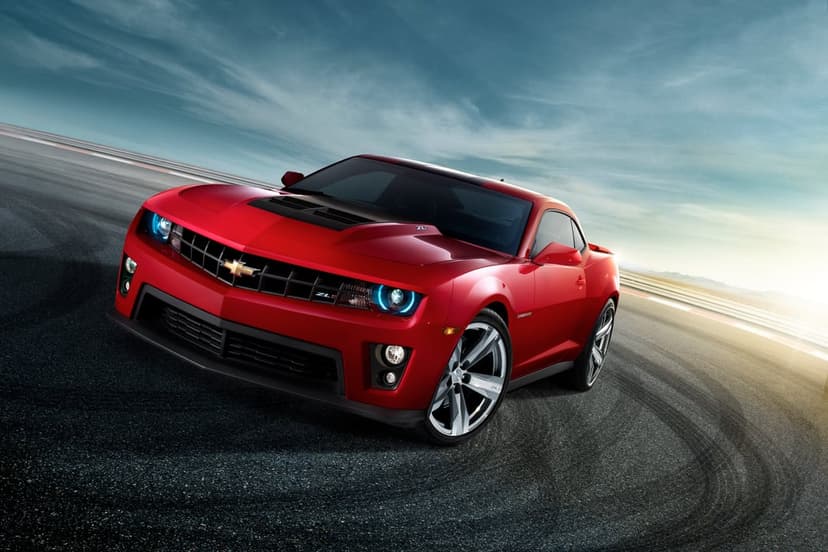Red Chevrolet Camaro ZL1 drifting on a racetrack with tire marks under a blue sky and golden sunlight.