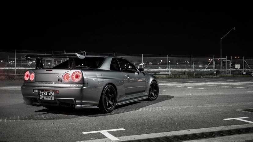 Rear view of a silver Nissan Skyline R34 GT-R with red tail lights glowing in a dark nighttime parking lot.
