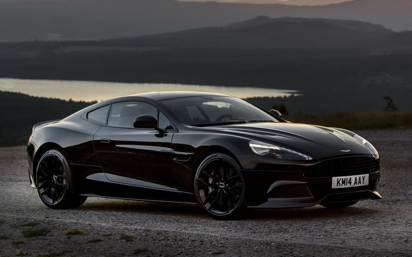 Black Aston Martin Vanquish parked on gravel with mountains and a lake in the background at sunset.