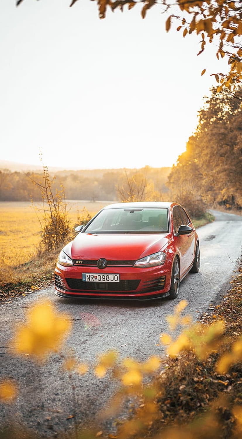 Red Volkswagen Golf GTI parked on an asphalt road surrounded by yellow and orange autumn trees in sunlight.